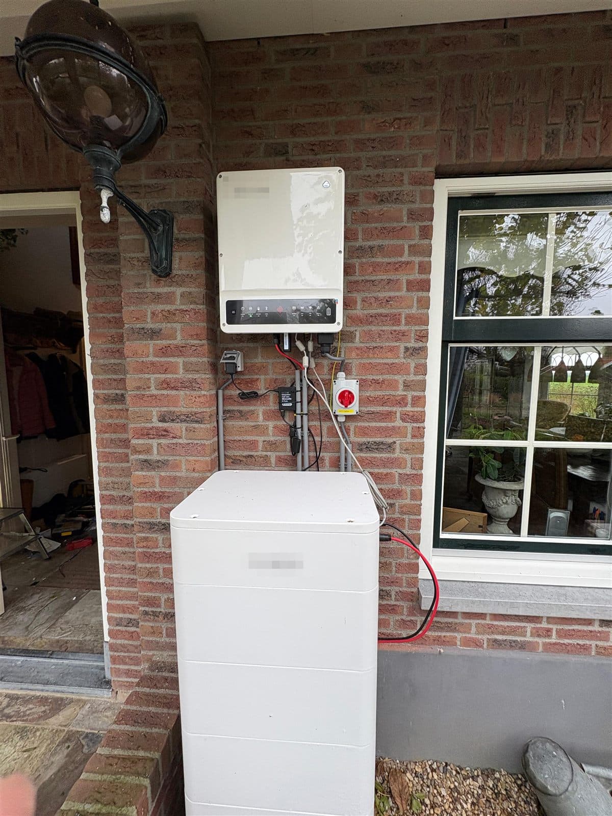 Thuisbatterij onbeschut buiten tegen een gevel, met losse kabels langs gevel en kozijn
